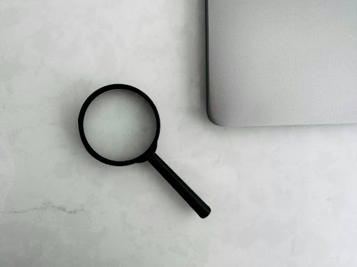 A magnifying glass rests beside a laptop, representing the periodic review of incident databases and shipment records that hazmat shippers should conduct to monitor regulatory exposure.